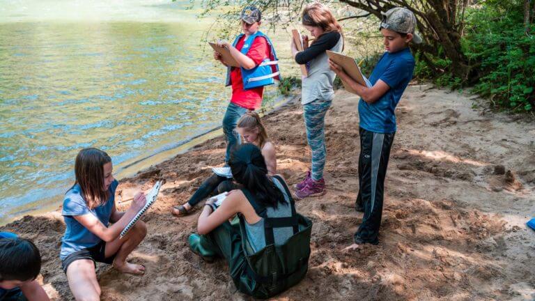 outdoor school students write in notebooks by river