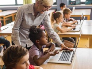 teacher leaning over shoulder of elementary student working on laptop at desk