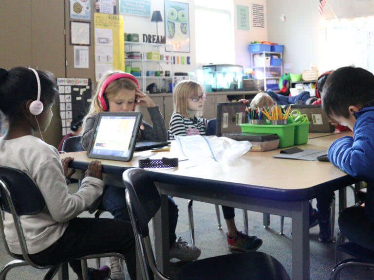 elementary students at a desk block, working on tablets