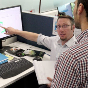 Male employee pointing to computer screen, looking up at standing male employee