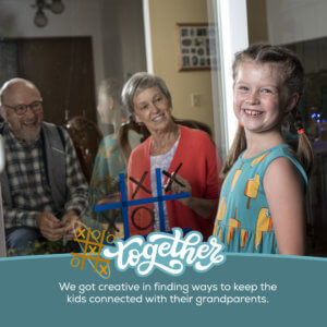 child laughing towards camera in front of a sliding glass door. Grandparents on the other side of the door, smiling at child, while they play tic tac toe on the glass