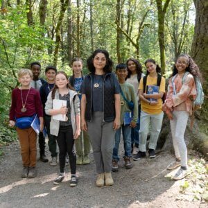 a teacher surrounded by elementary age students, all smiling at the camera, standing in a trail path in a forested area