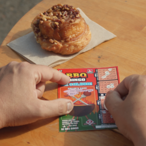 hand scratching a bingo-themed crossword, on a wooden table, with a sticky bun on a napkin to the left of the hand