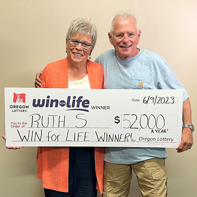 Ruth S and partner Ed, smiling in front of a beige background, holding a novelty oversize Oregon Lottery check