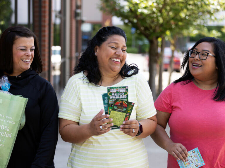 Three women, laughing in animated conversation are walking to the foreground. One woman holds a bag of groceries. The other two women are holding Scratch-its.