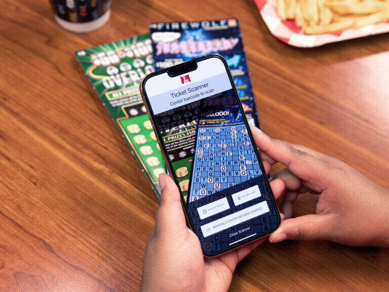 At a wooden restaurant table top, a pair of hands holds a phone over a pair of Scratch-its. The phone is open to the scanner function of the Oregon Lottery mobile app.