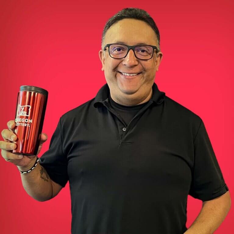 Video Lottery winner Gerardo A smiling and holding an Oregon Lottery travel mug.