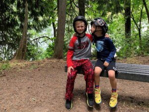 Two kids in helmets pose on a riverside bench.