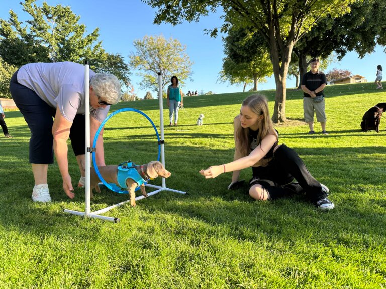 Two women help a small dog through an agility hoop
