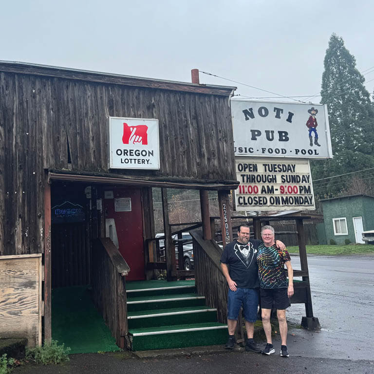 An exterior short of a semi-rural area, featuring a dark wood slated building. Next to the building is awhile sign with a cartoony illustration of a cowboy and the name 