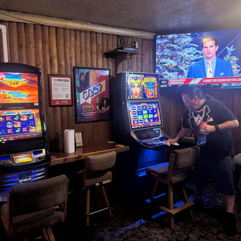 A Video Lottery play area with wood paneling, dark wood counters, and dark carpeted floor. You can see two VLTs, a large TV screen playing ESPN and a 