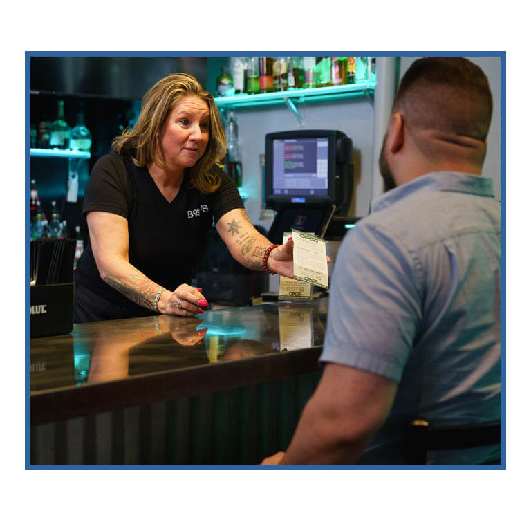 In a bar, we see over the shoulder of a man in a blue shirt sitting at the bar. Across from him is a female bartender in a black t-shirt. She is leaning over the bar offering him an OPGR brochure