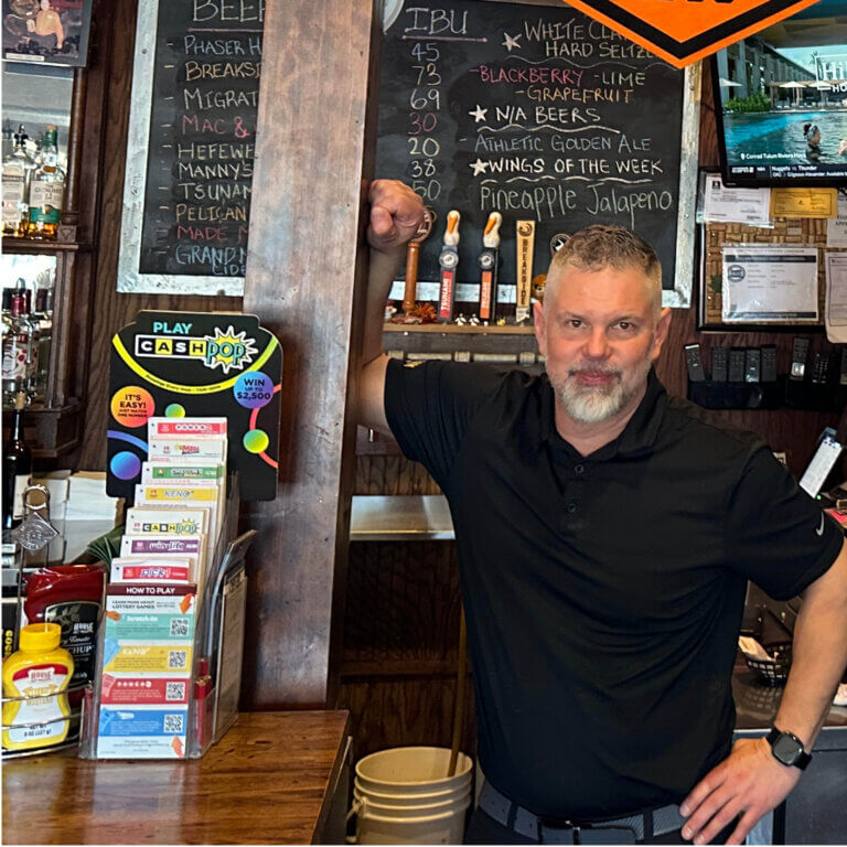A man with gray hair in a black polo shirt leans against the wooden pole of a bar area and smiles at the camera. Behind and around him is a crowded back bar space with draft bar pulls, a chalkboard menu sign, and an acrylic holder of lottery how-to-play brochures