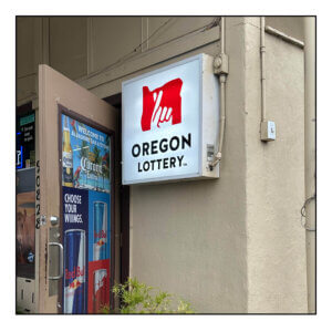 An exterior of a light brown stucco building. There is an open door plastered with posters of beers and energy drinks. Next to the door is an illuminated Oregon Lottery exterior sign.