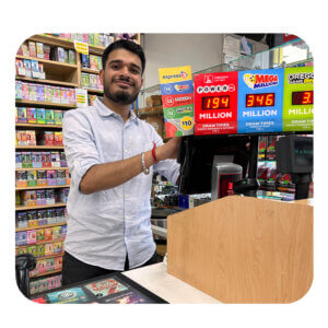 A smiling man of Southeast Asian descent with black hair stands behind the counter of a convenience store. Around him are Oregon Lottery jackpot signs and in front of him is a cash register.