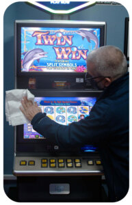 A man with short-cropped gray hair holds a white cloth and is wiping the screens of a Video Lottery terminal