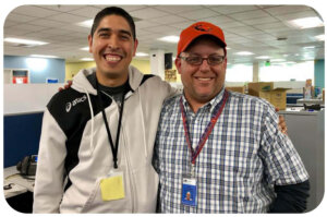 Inside of an office building, two men wearing casual clothes are smiling at the camera, with their arms draped across each other's shoulders in a friendly pose.