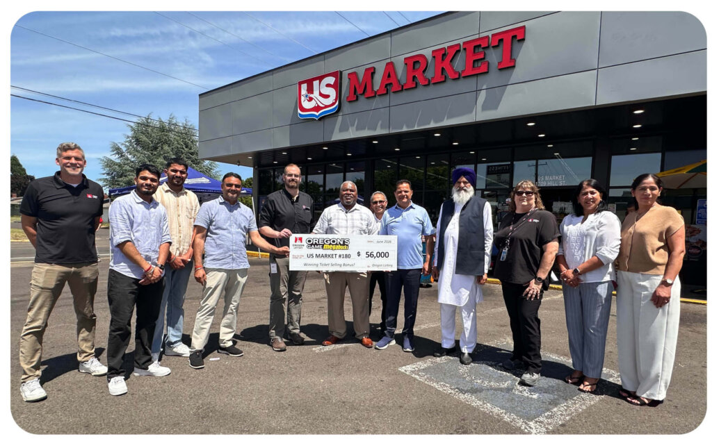 An external photo of US Market #180. Standing in front of the building are 12 people smiling at the camera. In the center, a man holds a novelty oversized check for Oregon Game's Megabucks, made out to US Market and labeled as a Winning Ticket Selling Bonus