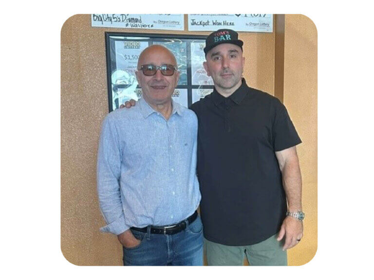 Two men are facing the viewer in semi-casual clothing. One is bald and wearing tinted glasses; the other is wearing a baseball cap that reads 