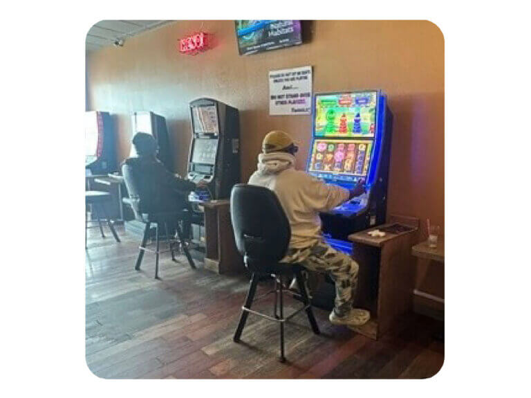 The interior of a Video Lottery play area with cappuccino-colored walls and dark wood plank flooring. There are four video lottery terminals. Each has a small wood side table next to it with a black bar stool in front. We can see two individuals in casual clothes with their back to the viewer seated in front of two VLTs